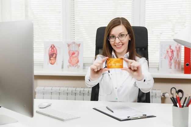 Giovane dottoressa sorridente seduta alla scrivania, con in mano una carta di credito, che lavora con documenti medici in un ufficio leggero in ospedale