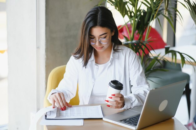 Giovane e affascinante libero professionista che utilizza il computer portatile per un lavoro a distanza mentre è seduto in un moderno bar interno bella donna caucasica che lavora su netbook durante la colazione mattutina nel bar caffetteria