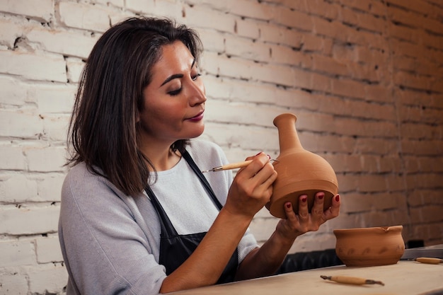 Giovane e attraente vasaio femminile che lavora in uno studio con un vaso di argilla marrone nel laboratorio artigianale che tiene in mano lo strumento e la tazza. donna d'affari nel negozio di ceramiche.