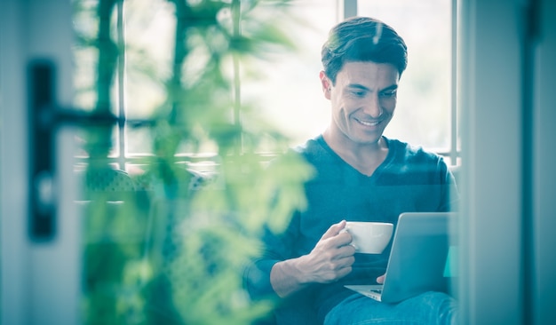 Giovane e bell'uomo d'affari caucasico che indossa un panno casual seduto felicemente sul divano, che tiene in mano una tazza e beve caffè e utilizza un computer portatile. Idea per lavorare a casa, lavoro in videochiamata.