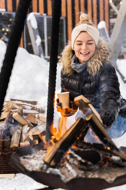 Giovane e bella donna che indossa un piumino che si scalda vicino al pozzo del fuoco durante la fredda giornata invernale