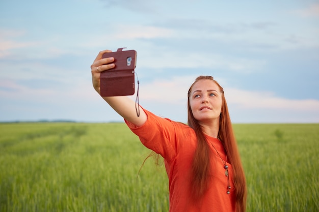 Giovane e bella donna in abito rosso e capelli rossi, prende un selfie al telefono nel campo di grano verde la sera