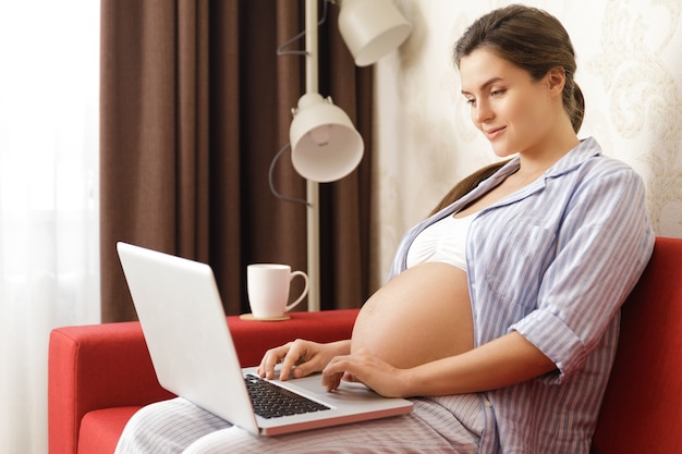 Giovane e bella donna incinta seduta con un computer portatile sul divano rosso