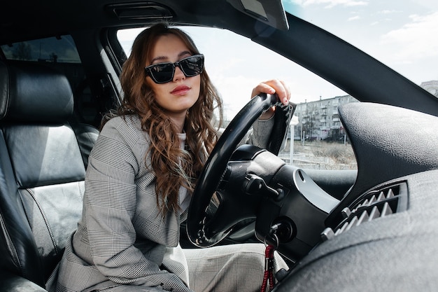 Giovane e bella ragazza alla guida di un'auto. Una ragazza alla moda in giacca e occhiali alla guida di un'auto nera.