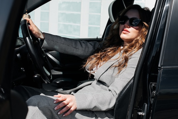 Giovane e bella ragazza alla guida di un'auto. Una ragazza alla moda in giacca e occhiali alla guida di un'auto nera.