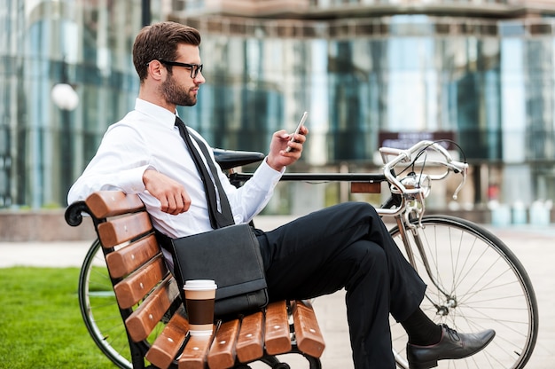 Giovane e di successo. Vista laterale di un bel giovane uomo d'affari che tiene il telefono cellulare mentre è seduto sulla panchina vicino alla sua bicicletta con un edificio per uffici sullo sfondo