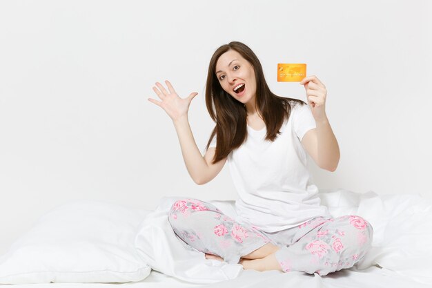 Giovane e divertente donna felice seduta a letto con un lenzuolo bianco, un cuscino, che si avvolge in una coperta sul muro bianco