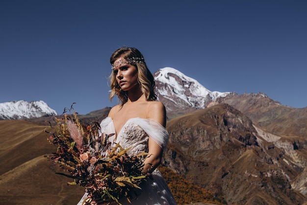 Giovane e donna attraente vestita in abito da sposa in stile boho in posa per una foto su sfondo di montagne trucco perfetto acconciatura e matrimonio floristica sposa felice in attesa dello sposo