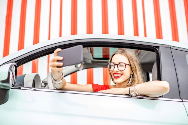 Giovane e donna felice che fa la foto del selfie con lo smartphone seduto in macchina sullo sfondo del muro rosso