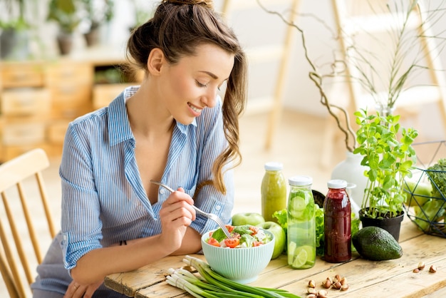 Giovane e donna felice che mangia insalata sana seduta sul tavolo con ingredienti freschi verdi al chiuso