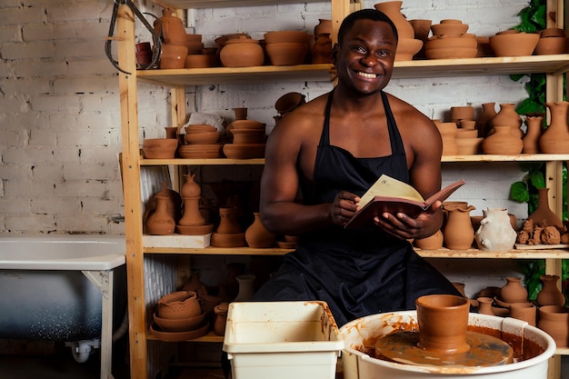 Giovane e fiducioso uomo afroamericano con un grembiule nero scolpisce un vasaio da un laboratorio di ceramica di argilla pentola di creta nella mano dell'artista latino