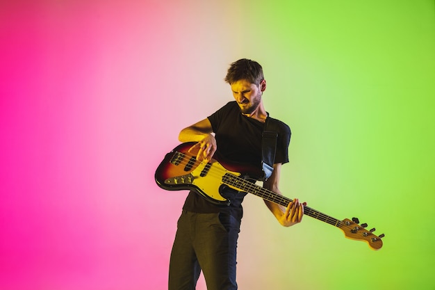 Giovane e gioioso musicista caucasico che suona il basso su sfondo sfumato per studio in luce al neon. Concetto di musica, hobby, festival. Ritratto colorato di artista moderno. Ispirato all'improvvisazione.