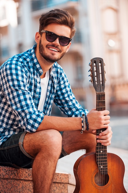 Giovane e spensierato. Giovane sorridente che tiene in mano la chitarra e guarda la telecamera mentre l'uomo è seduto all'aperto