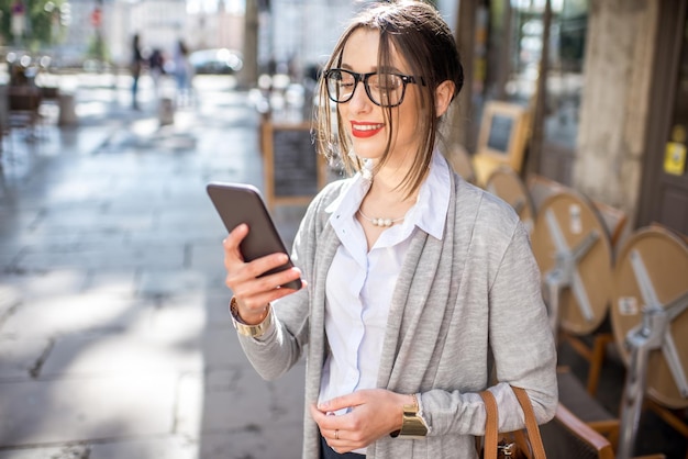 Giovane ed elegante imprenditrice in piedi con il telefono per strada con caffè nella città vecchia di Lione