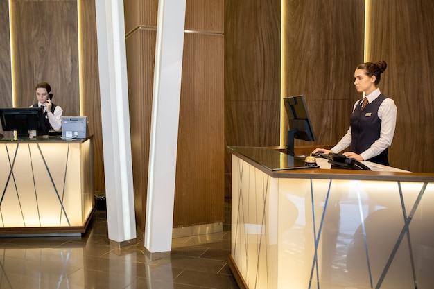 Giovane ed elegante receptionist femminile in uniforme in piedi al bancone e guardando lo schermo del computer mentre il suo collega risponde al telefono