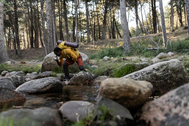 giovane escursionista afroamericano sta bevendo acqua da una tazza l'acqua di un fiume di montagna