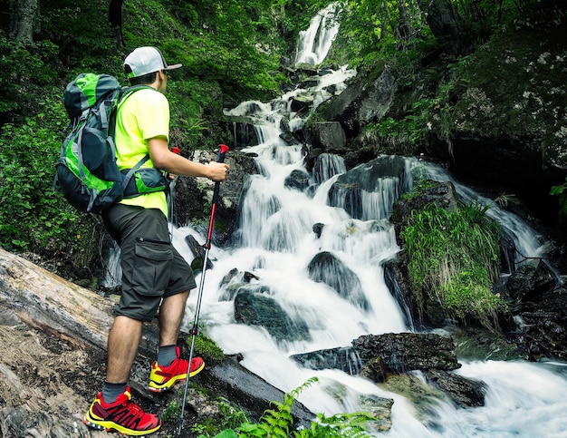 Giovane escursionista attivo con zaino e bastoncini da trekking guardando la cascata nella foresta