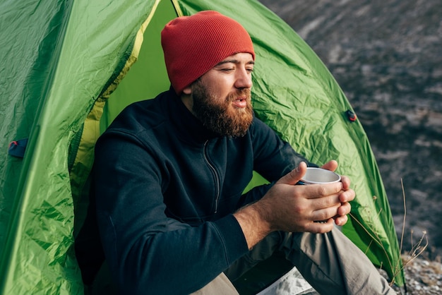 Giovane esploratore che beve bevande calde in montagna seduto vicino al falò rilassante dopo il trekking Uomo viaggiatore con cappello rosso che tiene una tazza di tè dopo l'escursionismo Viaggiatore
