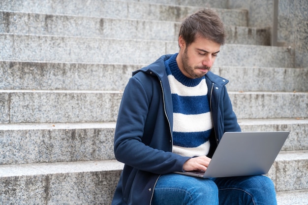 giovane espressione seria, uomo libero professionista che lavora con il computer portatile all'aperto seduto a gradini, in uno scenario urbano.