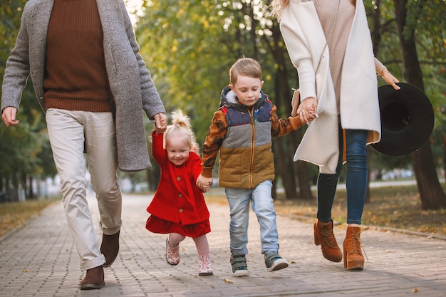 Giovane famiglia cammina lungo un vicolo del marciapiede in un parco. Lui indossa un cappotto grigio e occhiali neri, lei è in bianco, la ragazza è bionda in rosso.