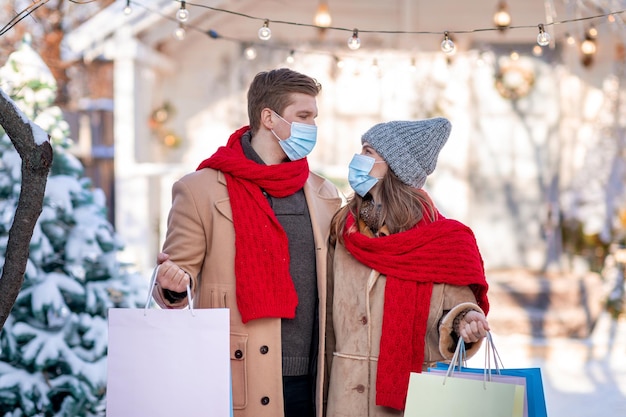 Giovane famiglia che fa shopping durante la pandemia di covid-19, uomo e donna in abiti caldi che indossano maschere protettive, tengono acquisti e si guardano, tornano a casa, concetto di saldi invernali