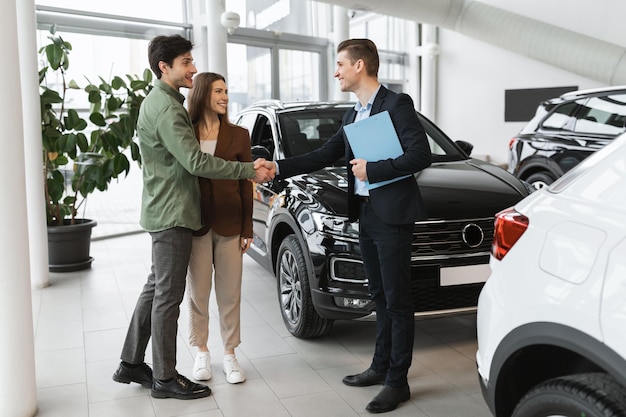 Giovane famiglia che fa un affare per l'acquisto o l'affitto di un'automobile moderna che stringe la mano al venditore di auto all'auto