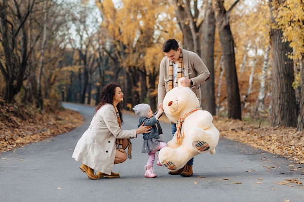 Giovane famiglia con la piccola figlia al giocattolo del grande orso del presente della strada del parco di autunno