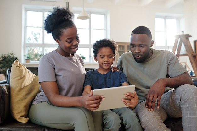 Giovane famiglia contemporanea di padre, madre e il loro simpatico figlioletto seduti sul divano in soggiorno e guardando film online insieme