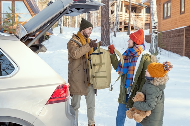 Giovane famiglia di tre persone in abiti invernali caldi che tirano fuori il bagagliaio dal bagagliaio mentre sono in piedi sul retro dell'auto durante i viaggi invernali