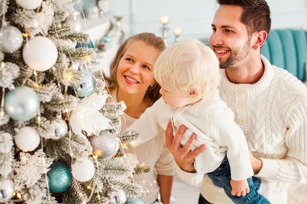 Giovane famiglia felice con bambino vicino albero di Natale decorato Capodanno divertimento con la famiglia a casa