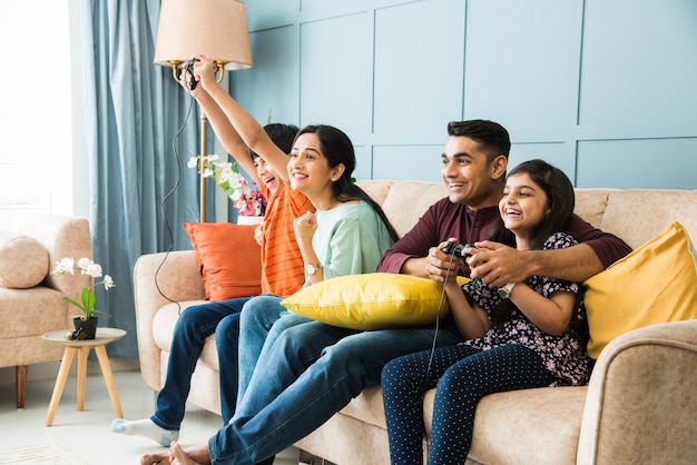 Giovane famiglia indiana di quattro persone che giocano ai videogiochi utilizzando il controller o il joystick mentre sono seduti sul divano