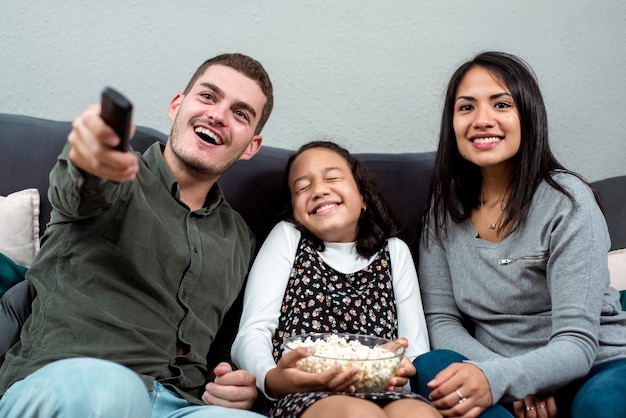 Giovane famiglia interrazziale che guarda a bocca aperta la TV dal divano. Il giovane telecomando del padre caucasico e la figlia tengono il popcorn.