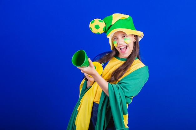 Giovane fan di calcio del Brasile vestito con cappello e bandiera che fa rumore con il clacson