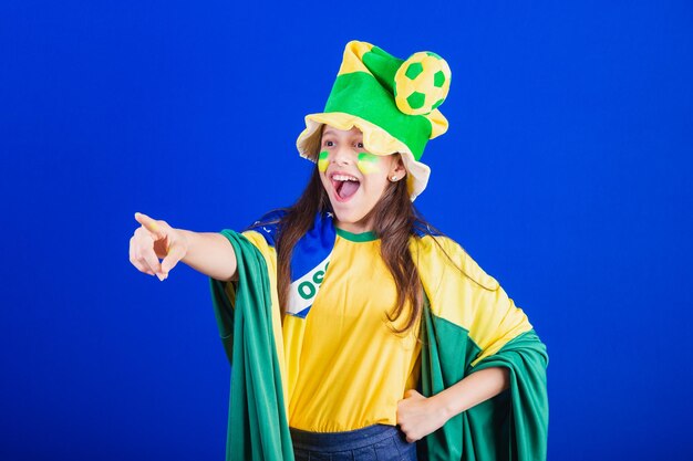 Giovane fan di calcio del Brasile vestito con cappello e bandiera che puntano a qualcosa di lontano