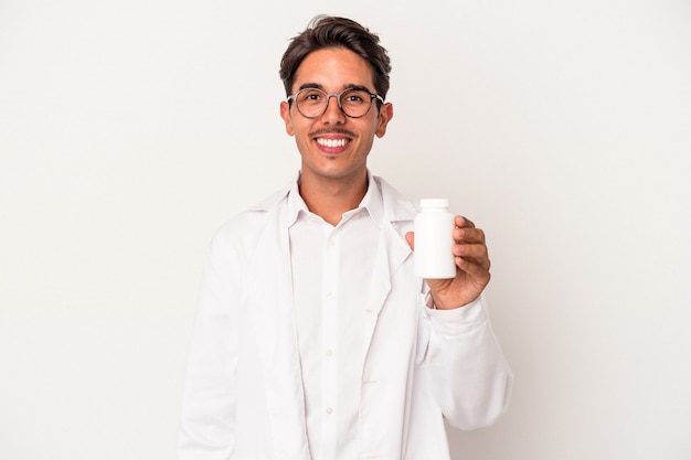 Giovane farmacista uomo di razza mista che tiene pillole isolate su sfondo bianco felice, sorridente e allegro.