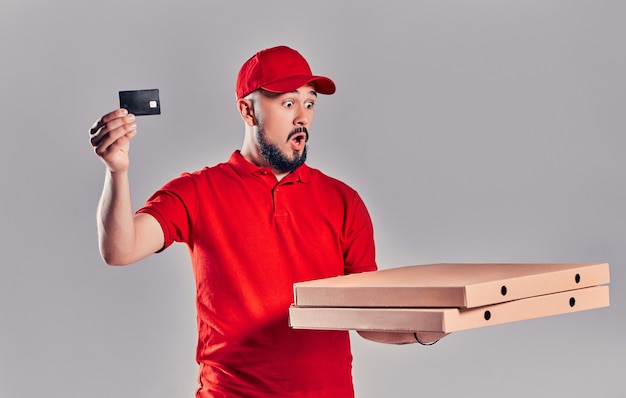 Giovane fattorino barbuto in uniforme rossa con pizza e carta di credito isolato su sfondo grigio.