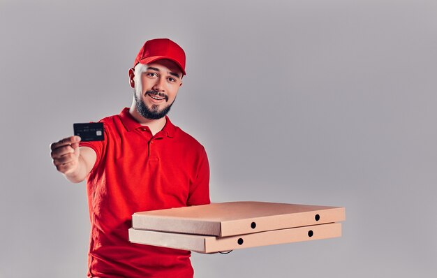 Giovane fattorino barbuto in uniforme rossa con pizza e carta di credito isolato su sfondo grigio.