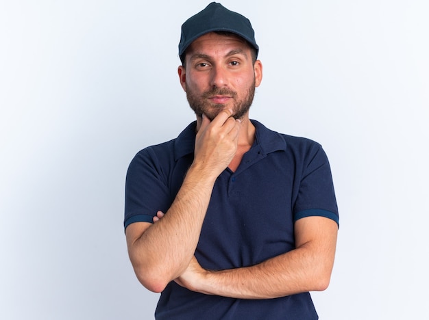 Giovane fattorino caucasico premuroso in uniforme blu e berretto che tiene la mano sul mento