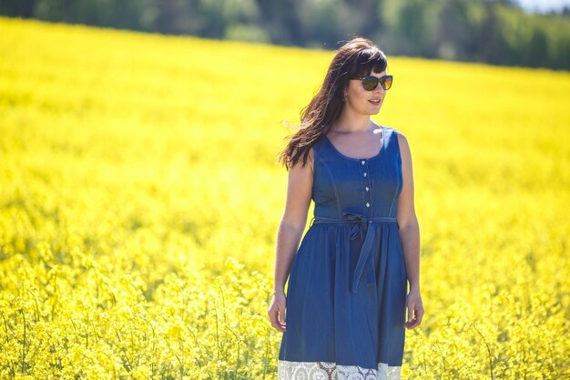 Giovane felice bella ragazza modello di grandi dimensioni in abito blu con occhiali da sole sul campo di colza in fiore in estate