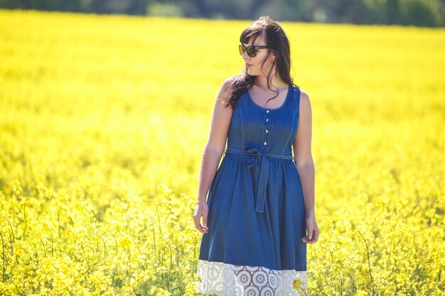 Giovane felice bella ragazza modello di grandi dimensioni in abito blu in occhiali da sole sul campo di colza in fiore in estate