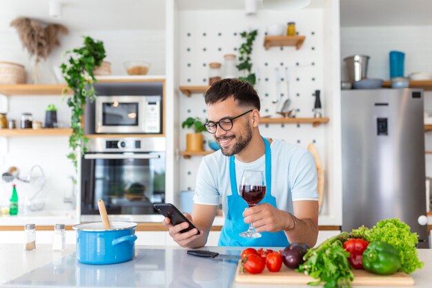 Giovane felice che prepara una cena romantica alla ricerca di ricette di verdure, menu dietetici, app di libri di cucina usando lo smartphone, marito sorridente che tiene in mano il telefono, cucina, cibo vegano sano, insalata tagliata nell'interno della cucina.