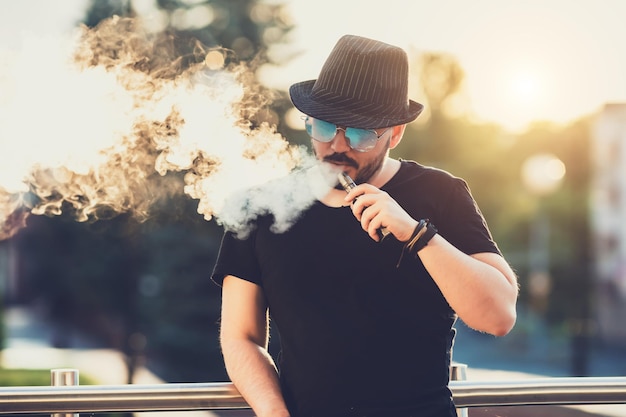 Giovane felice moderno con cappello con una barba divertente Vaporizzatori Sullo sfondo il tramonto serale