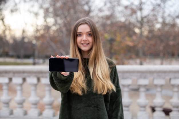 Giovane femmina adorabile in attrezzatura alla moda che sorride e che esamina macchina fotografica mentre stando nel parco e dimostrando smartphone moderno con lo schermo in bianco