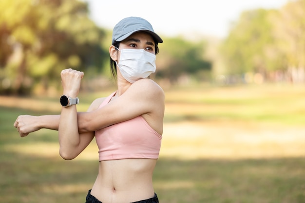 Giovane femmina adulta che indossa la maschera protettiva durante lo stretching muscolare nel parco all'aperto