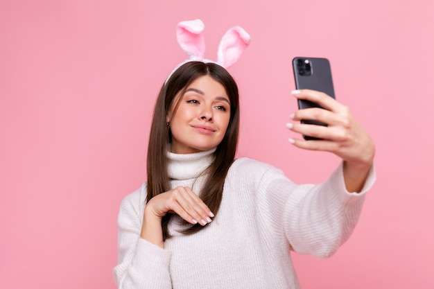 Giovane femmina adulta con orecchie da coniglio con un sorriso attraente e giocoso che si fa selfie o fa una videochiamata indossando un maglione bianco in stile casual Colpo in studio al coperto isolato su sfondo rosa