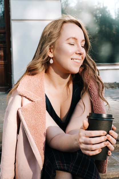 Giovane femmina adulta sorridente in pelliccia sintetica che si siede all'aperto con la tazza della bevanda calda in sue mani