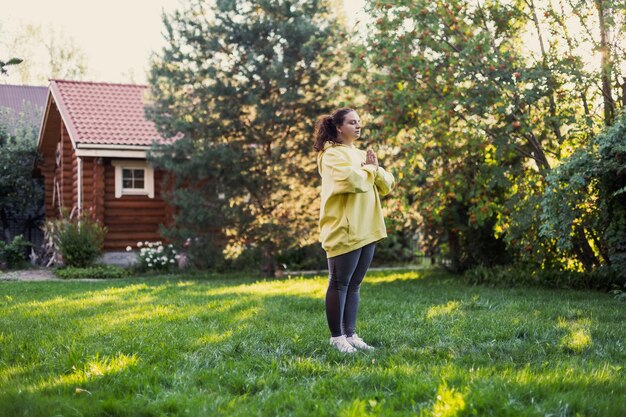 Giovane femmina adulta su erba verde che media fuori in abbigliamento sportivo sul cortile pieno di luce solare con casa di campagna e alberi ad alto fusto sullo sfondo Corpo positivo