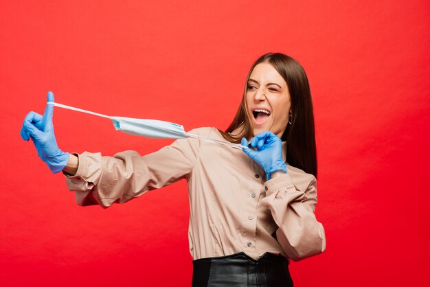 Giovane femmina allegra e allegra che gioca con la mascherina medica protettiva e guanti blu su priorità bassa rossa. Ritratto del primo piano Fine della quarantena del coronavirus, isolamento. Covid-2019