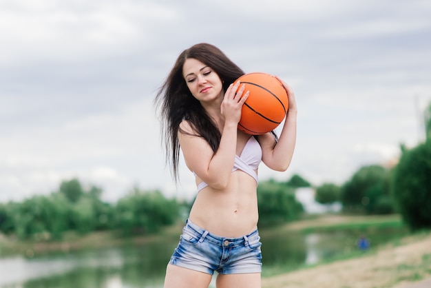 Giovane femmina atletica, in un top e pantaloni della tuta, che gioca con la palla sul campo da basket all'aperto.