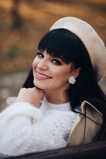 Giovane femmina caucasica allegra con capelli scuri lunghi in camicetta bianca, pantaloni grigi, cappello bianco e cappotto grigio con borsa marrone nella foresta